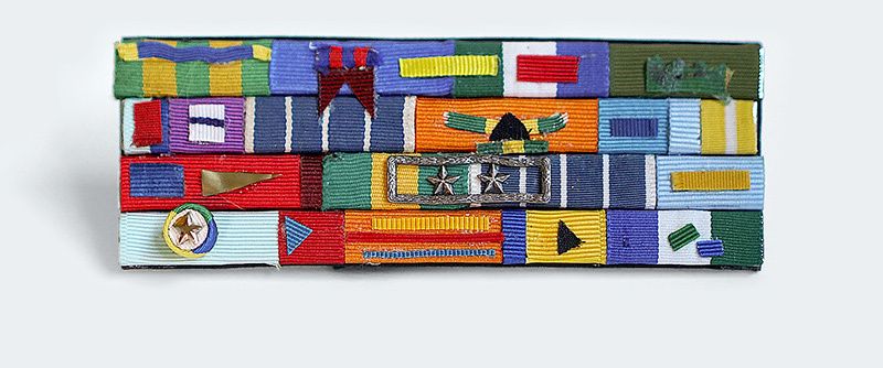 Pequeno broche que faz alusão à broches de condecoração militar, é composto de pequenas faixas de fitas de tecido coloridos, organizadas em formas geométricas diversas, coladas em placas de metal.