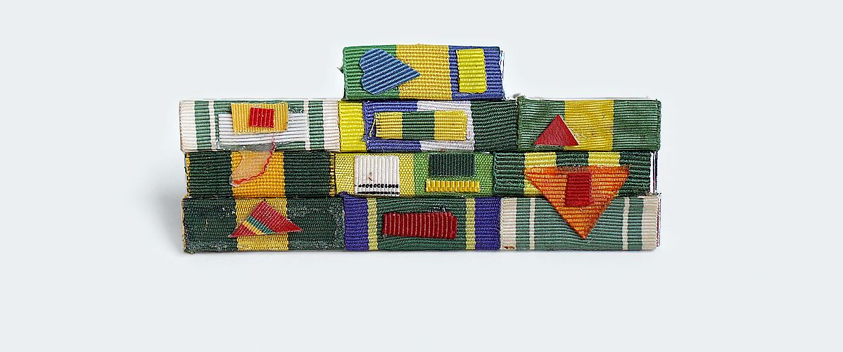 Pequeno broche que faz alusão à broches de condecoração militar, é composto de pequenas faixas de fitas de tecido coloridos, organizadas em formas geométricas diversas, coladas em placas de metal.